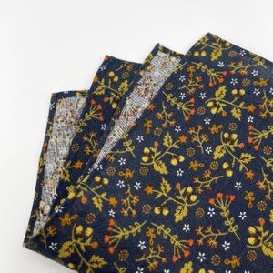 May include: Folded navy blue fabric with a floral pattern. The design features small white flowers, orange berries, and yellow-green leaves. The fabric is layered, revealing a glimpse of a lighter, patterned interior.