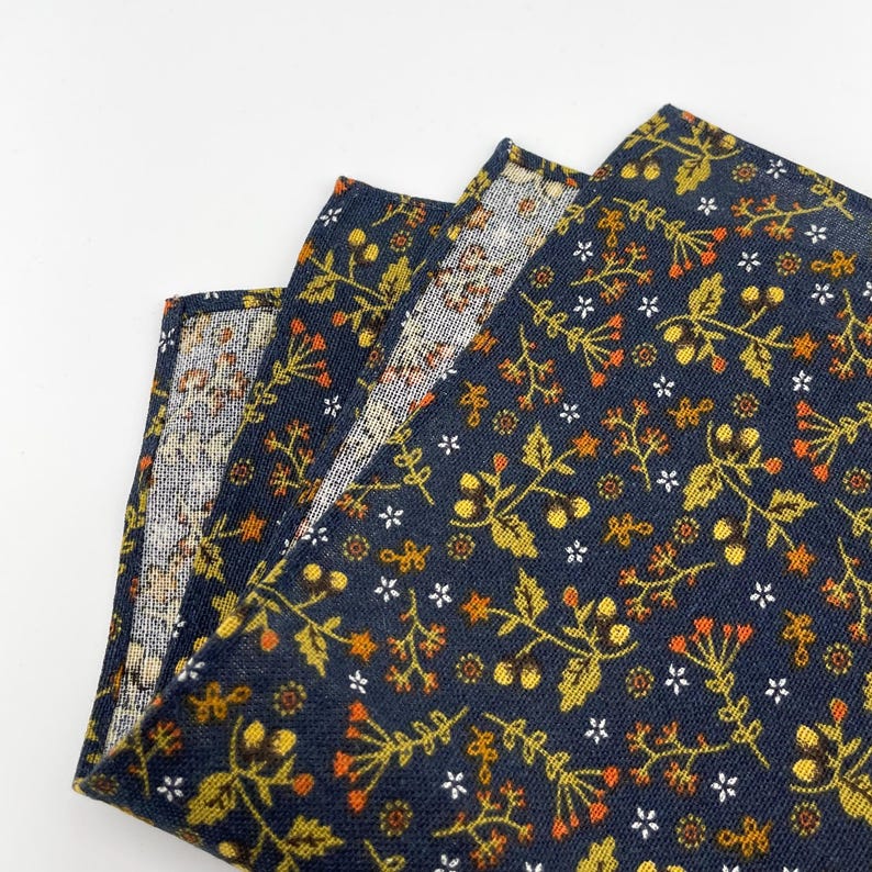 May include: Folded navy blue fabric with a floral pattern. The design features small white flowers, orange berries, and yellow-green leaves. The fabric is layered, revealing a glimpse of a lighter, patterned interior.