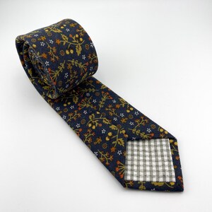 May include: A navy blue tie with a floral pattern in shades of gold, orange, and white. The tie is rolled up, showing the patterned fabric and the gingham lining on the back.