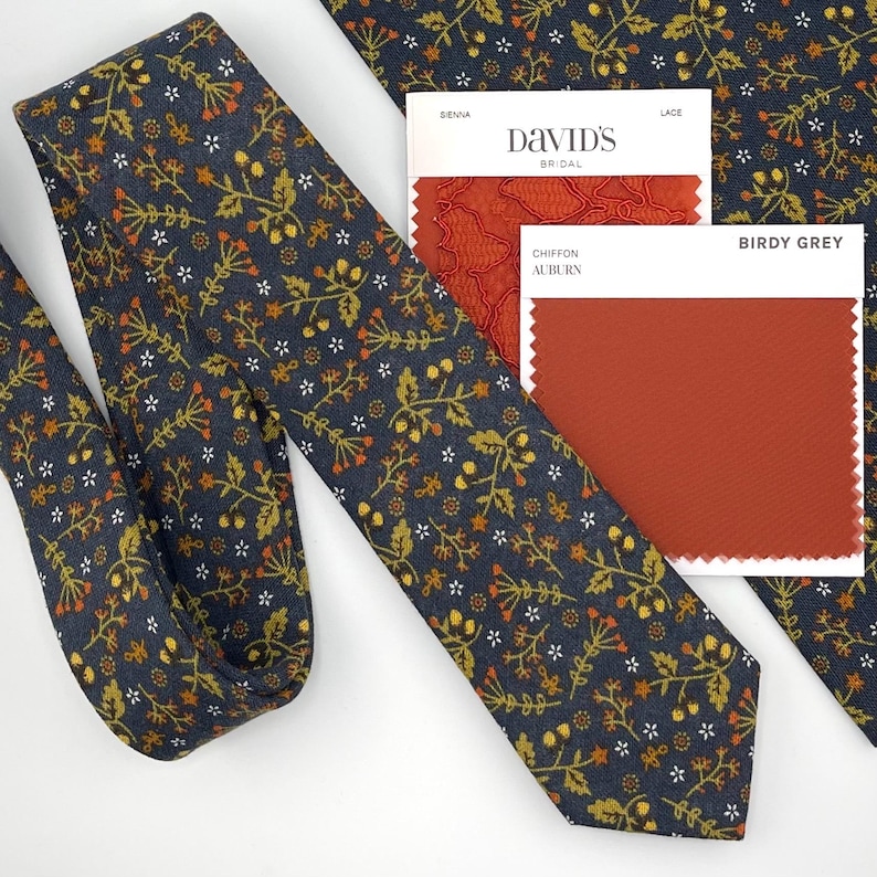 May include: A navy blue tie with a floral pattern in gold, orange, and white. The tie is next to a matching pocket square and color swatches from David's Bridal and Birdy Grey, including Sienna, Lace, and Chiffon Auburn.