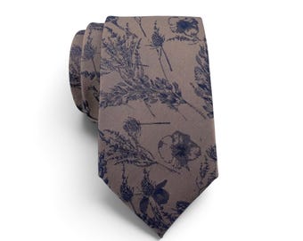 Può includere: Una cravatta color tortora con una stampa botanica blu navy scuro raffigurante vari fiori di campo, erbe e capolini. La cravatta è piegata nella parte superiore, mostrando la trama del tessuto e il motivo ripetitivo ispirato alla natura.