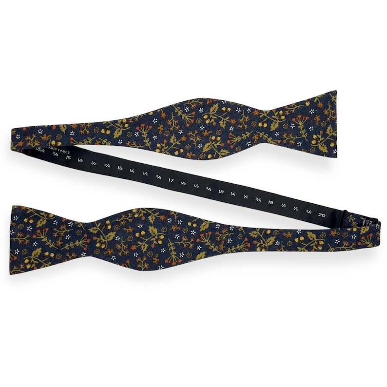 May include: A navy blue adjustable bow tie with a floral pattern in gold, orange, and white. The bow tie is open, showing the adjustable strap with size markings from 14.5 to 20 inches.