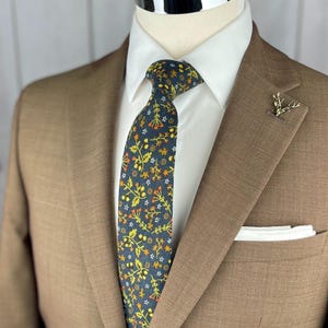 May include: A brown suit with a white dress shirt and a patterned tie. The tie features a dark blue background with yellow, orange, and white floral designs. A gold-colored lapel pin and a white pocket square complete the look.