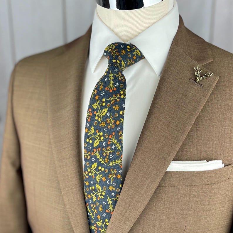 May include: A brown suit with a white dress shirt and a patterned tie. The tie features a dark blue background with yellow, orange, and white floral designs. A gold-colored lapel pin and a white pocket square complete the look.