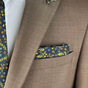 May include: A brown suit jacket with a white shirt underneath. A navy blue tie with a floral pattern is visible, along with a matching pocket square. The pocket square and tie feature yellow and white floral designs.