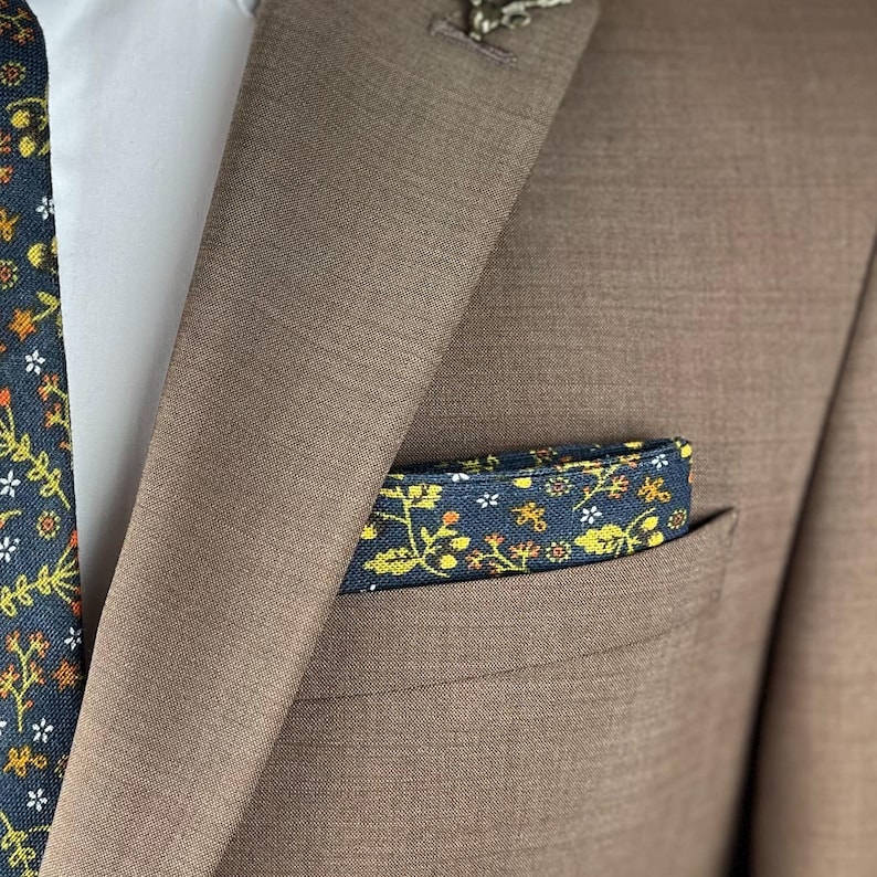 May include: A brown suit jacket with a white shirt underneath. A navy blue tie with a floral pattern is visible, along with a matching pocket square. The pocket square and tie feature yellow and white floral designs.
