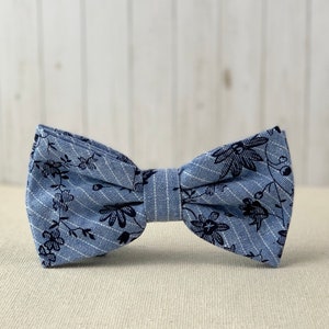 May include: A blue bow tie with a black floral pattern. The bow tie is made of a textured fabric.