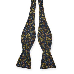 May include: A navy blue bow tie with a repeating floral pattern in shades of gold, orange, and white. The bow tie is made of a textured fabric and is designed to be self-tied. The overall design is a classic and elegant accessory.