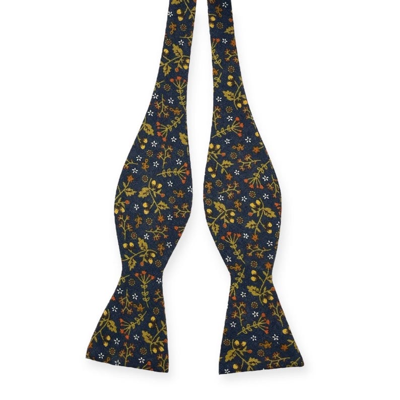 May include: A navy blue bow tie with a repeating floral pattern in shades of gold, orange, and white. The bow tie is made of a textured fabric and is designed to be self-tied. The overall design is a classic and elegant accessory.