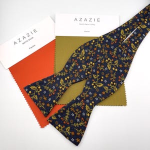 May include: A navy blue bow tie with a floral pattern in gold, orange, and white, next to fabric swatches. The swatches are paprika orange and olive green. The bow tie is made of stretch satin and the fabric swatches are matte satin.
