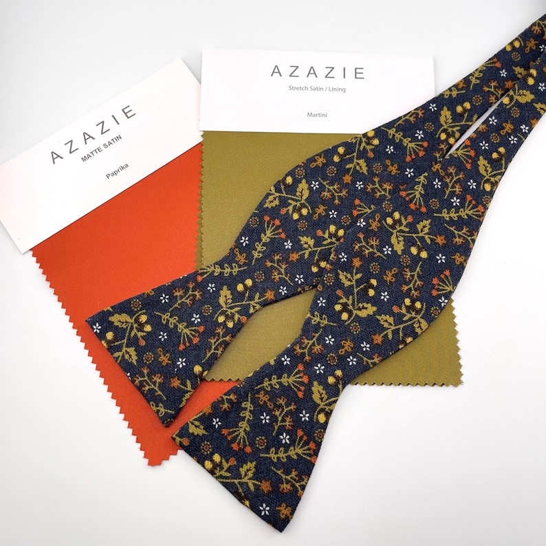 May include: A navy blue bow tie with a floral pattern in gold, orange, and white, next to fabric swatches. The swatches are paprika orange and olive green. The bow tie is made of stretch satin and the fabric swatches are matte satin.