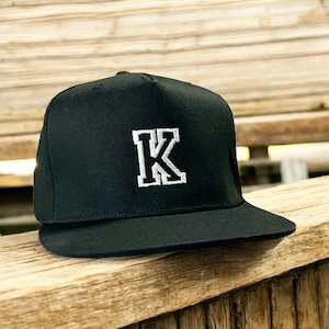 Könnte beinhalten: Eine schwarze Baseballkappe mit einem weißen gestickten Buchstaben 'K' auf der Vorderseite.