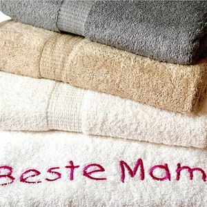 Könnte beinhalten: Ein Stapel aus vier flauschigen Badetüchern in Grau-, Beige- und Weißtönen. Das untere weiße Handtuch ist mit den Worten "Beste Mama" in leuchtendem Pink bestickt. Die Handtücher sind gefaltet und gestapelt.