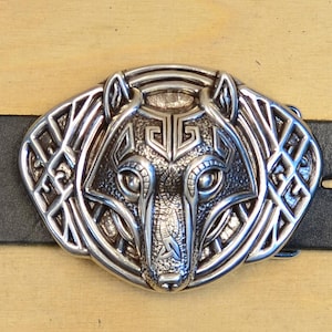 May include: Silver-colored belt buckle with a wolf design, attached to a black leather belt. The buckle features intricate detailing of a wolf's head. A brown leather loop with the number "1" is visible.