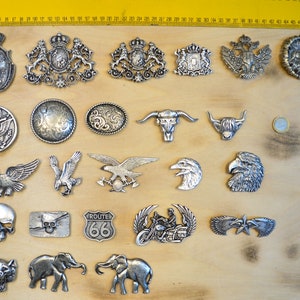Puede incluir: Una colección de hebillas de cinturón de metal plateado con varios diseños, incluyendo águilas, calaveras y escudos heráldicos. Las hebillas se muestran sobre una superficie de madera con una regla amarilla en el fondo. La colección incluye una hebilla de la Ruta 66 y un diseño de motocicleta.