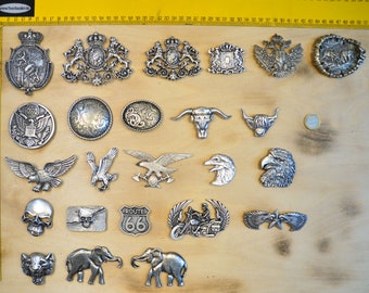 May include: A collection of silver-colored metal belt buckles with various designs, including eagles, skulls, and heraldic crests. The buckles are displayed on a wooden surface with a yellow ruler in the background. The collection includes a Route 66 buckle and a motorcycle design.