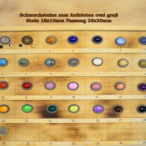 Könnte beinhalten: Eine Holzkassette mit 35 ovalen, bunten Glascabochons in silbernen Fassungen. Die Cabochons sind in Reihen angeordnet und mit den Zahlen 1 bis 35 beschriftet. Der Text "Schmucksteine zum Aufnieten oval groß Stein 18x10mm Fassung 25x20mm" ist oben auf der Kassette aufgedruckt.