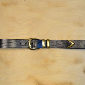 May include: A black leather belt with a brass buckle and accents. The belt features a classic design with a rectangular buckle and two brass loops. The belt has a brass arrow-shaped detail near the end.