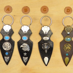 May include: Four leather keychains with different designs. Each keychain features a silver or brass ring, a leather arrowhead-shaped charm, and a decorative metal design. The designs include a Native American profile, an eagle, a wolf, and a horse.