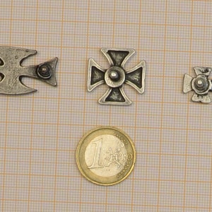 May include: Four silver metal crosses of varying sizes, with a single euro coin for scale. The crosses have a simple design with a raised center and four points.