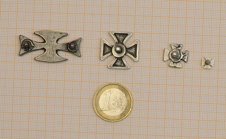 May include: Four silver metal crosses of varying sizes, with a single euro coin for scale. The crosses have a simple design with a raised center and four points.