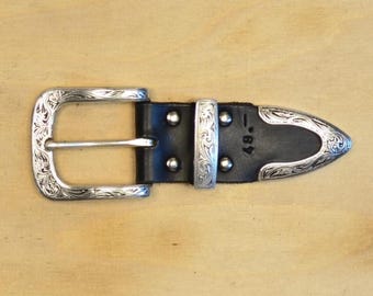 May include: A black leather belt end featuring a silver-toned metal buckle and a pointed tip, both decorated with intricate engraved scrollwork patterns. The leather strap is secured with four silver-toned rivets and includes the number 49 stamped.