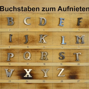 Può includere: Un set di 28 lettere metalliche per ribadire, disposte in ordine alfabetico su una tavola di legno. Le lettere sono di colore argento e hanno una finitura leggermente testurizzata.