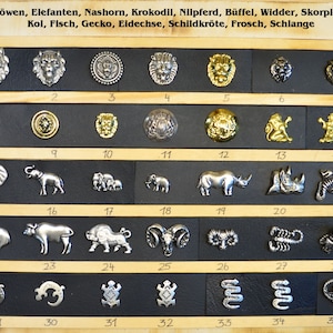 Puede incluir: Una muestra de 35 botones de metal plateado y dorado con diseños de animales y naturaleza. Los botones están dispuestos en filas sobre un fondo negro con números blancos que indican su posición.