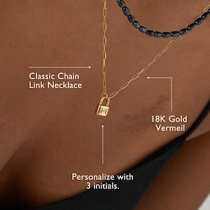 Może przedstawiać: Zestaw naszyjników warstwowych ze złotym naszyjnikiem z łańcuszkiem i zawieszką w kształcie kłódki, złotym łańcuszkiem i naszyjnikiem z ciemnoniebieskich koralików. Zawieszka w kształcie kłódki jest spersonalizowana inicjałami. Tekst brzmi "Classic Chain Link Necklace" i "18K Gold Vermeil".