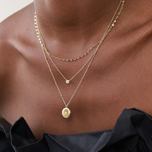 Puede incluir: Un collar de oro de varios niveles con un pequeño colgante redondo y un colgante ovalado más grande. El collar lo lleva una persona de piel oscura.