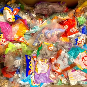 May include: A cardboard box filled with numerous sealed plastic bags containing various Ty Beanie Babies. The bags are colorful, with some featuring McDonald's branding. The toys are in various shapes and colors.