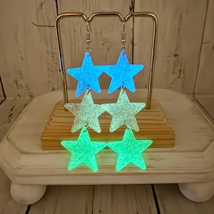 May include: A pair of dangling earrings featuring three stacked star-shaped charms each. The top stars are bright blue, the middle stars are a pale translucent white, and the bottom stars are a vibrant green. All stars have a glittery finish.