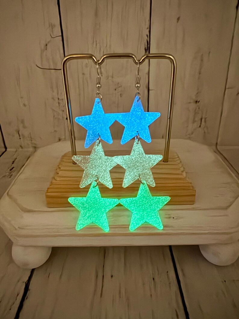 May include: A pair of dangling earrings featuring three stacked star-shaped charms each. The top stars are bright blue, the middle stars are a pale translucent white, and the bottom stars are a vibrant green. All stars have a glittery finish.