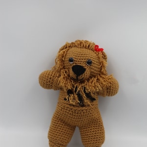 May include: A brown crocheted lion stuffed animal with black eyes and a black nose. The lion has a red bow on its head and is wearing a brown crocheted outfit.
