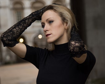 May include: A woman wearing a black turtleneck sweater and black lace gloves. The gloves extend to the elbows. The woman has blonde hair and is looking to the side. The background is a blurred outdoor scene.