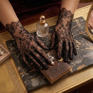 May include: Black lace gloves with intricate floral patterns are displayed on a marble surface. A vintage perfume bottle, a small antique book, and a jewelry box with pearl earrings are also visible. A small mirror reflects a person's face.
