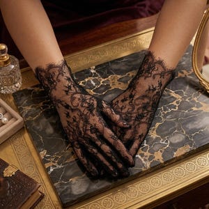 May include: Black lace gloves with intricate floral patterns. The gloves are displayed on a marble surface, with a gold-framed mirror and jewelry box in the background. The gloves have a delicate, scalloped edge.