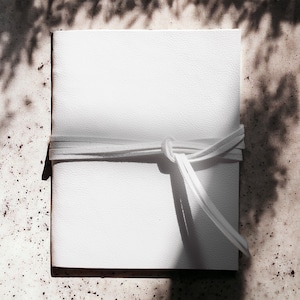 May include: A white leather journal with a white leather strap tied around it. The journal is sitting on a textured surface.
