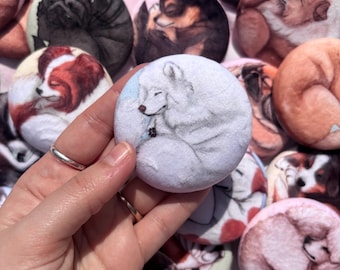 May include: A collection of round, fabric-covered buttons featuring sleeping dog illustrations. The central button shows a white dog curled up, with others depicting various breeds in shades of brown, black, and red. The buttons are held by a hand.