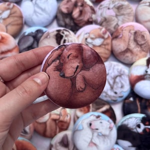 Puede incluir: Una colección de pines redondos ilustrados con perros. Uno de los pines muestra un perro salchicha durmiendo en tonos marrón y beige, con un borde rosa claro. Otros pines representan varias razas de perros en estilos de acuarela similares.