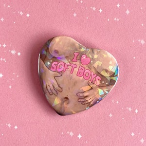 May include: A heart-shaped pin with a holographic finish featuring a cartoon illustration of a person's torso with the text "I ♥ SOFT BOYS" in pink.