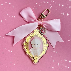 May include: A pink keychain with a gold clasp and a pink satin bow. The charm features a portrait of a white dog in a gold ornate frame, with a pink heart-shaped charm. The background is pink with white sparkles.