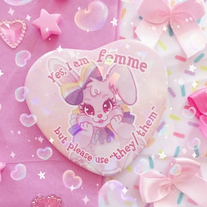 Puede incluir: Un botón rosa en forma de corazón con un conejito de dibujos animados que lleva un lazo y una banda de colores arcoíris. El texto en el botón dice "Sí, soy femme pero por favor usa "ellos/ellas"."