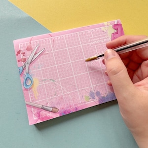 Puede incluir: Una estera de corte autocicatrizante rosa con un patrón de cuadrícula, salpicaduras de pintura e ilustraciones de tijeras, un pincel y una regla. El texto "crafters essential cutting mat" está impreso en la estera.