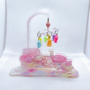 Könnte beinhalten: Ein Baby-Mobile aus rosa und klarem Acryl mit bunten Bären- und Tieranhängern. Die Basis zeigt einen schlafenden Welpen, einen Teddybären und ein Kaninchen. Das Design verwendet eine Pastellfarbpalette.