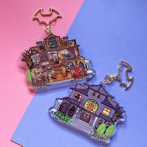 Pode incluir: Dois chaveiros de acrílico com um design de casa assombrada. Um chaveiro mostra o interior da casa com figuras de desenhos animados. O outro é uma casa roxa com uma janela de vitral. Ambos têm um fecho dourado em forma de morcego.