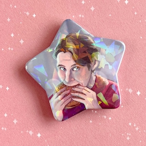 Può includere: Un bottone a forma di stella olografico con un ritratto di una persona con i capelli castani che morde un hamburger. La persona indossa una camicia rossa.