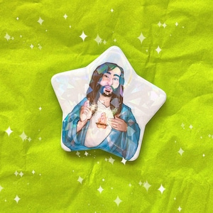 May include: A white star-shaped pin with a colorful illustration of a person with long brown hair and a beard wearing a blue robe with a white shirt underneath. The person is holding their hands together in front of their chest.