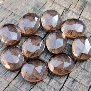 May include: Ten faceted oval-shaped smoky quartz gemstones in various shades of brown. The stones are arranged in a circle on a wooden surface.