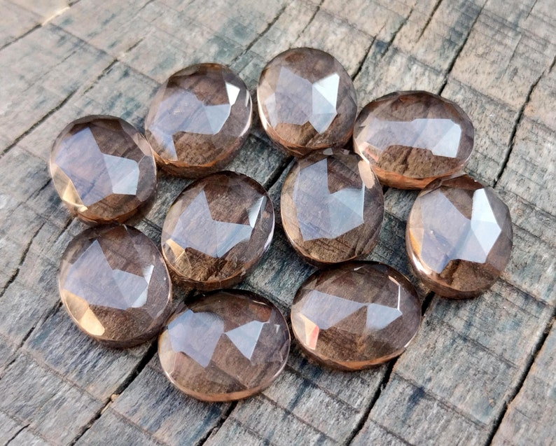 May include: Ten faceted oval-shaped smoky quartz gemstones in various shades of brown. The stones are arranged in a circle on a wooden surface.